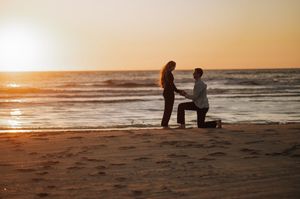 Man kneeling on one knee proposing to a woman on a sandy beach at sunset, with golden light reflecting on the ocean waves.