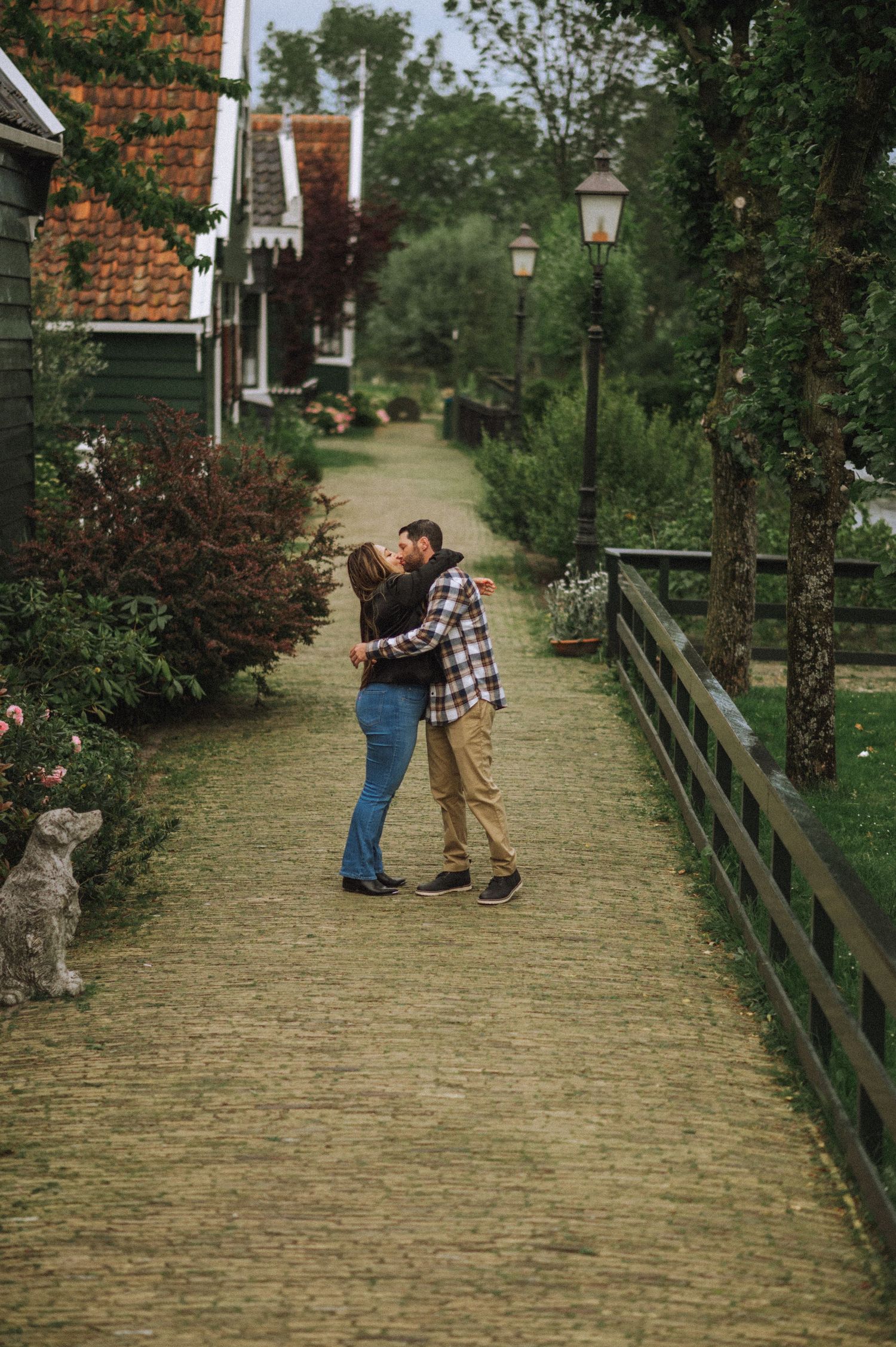 Couple kissing on a brick-paved path lined with greenery, lamp posts, and a wooden fence near traditional houses with red tile roofs.