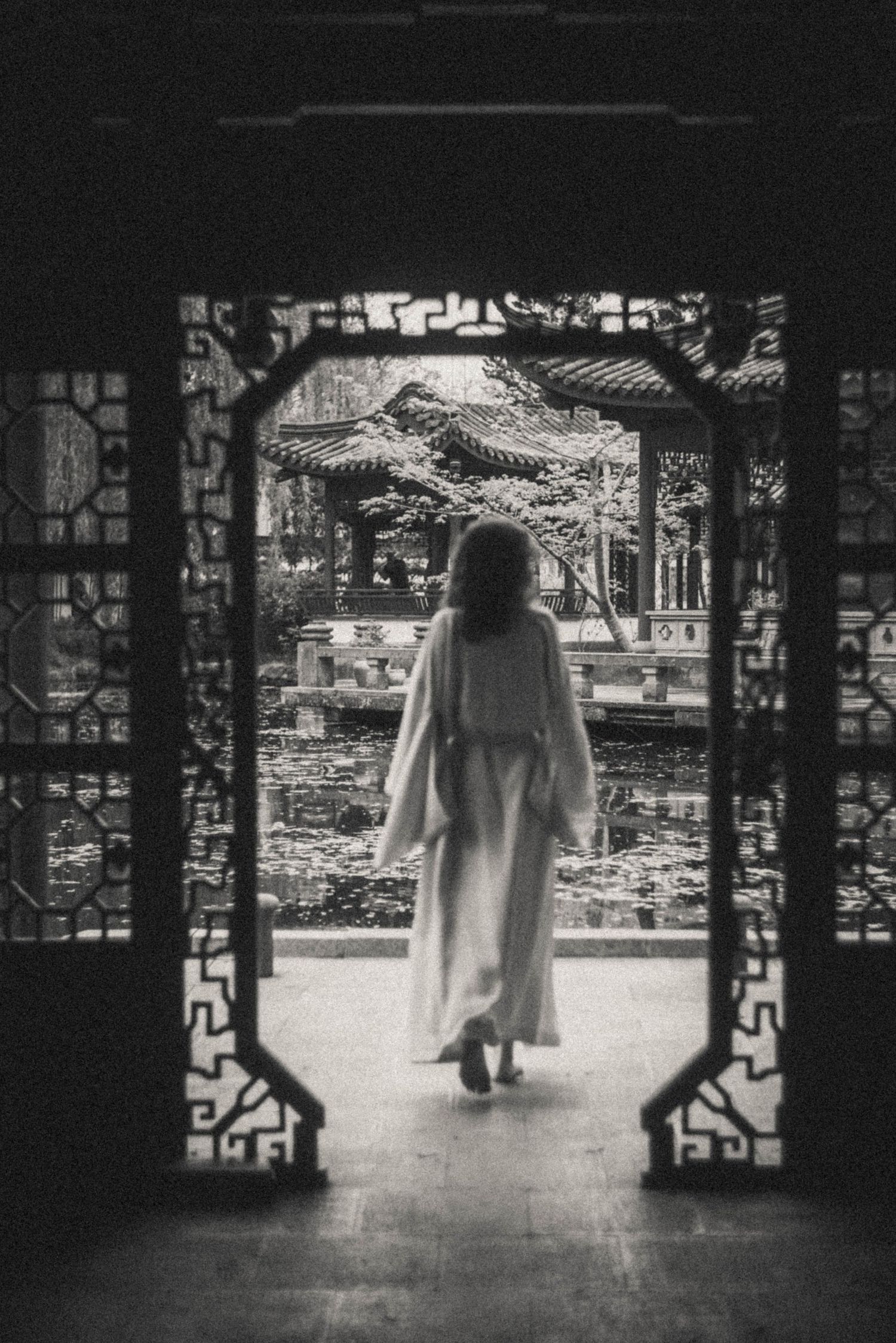 Woman in a long flowing robe walks through an ornate carved doorway toward a classical Chinese garden pavilion and pond in black and white.
