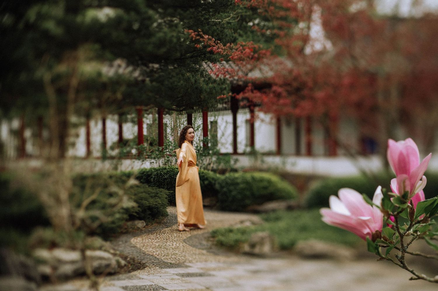 Woman in a flowing yellow dress walks along a stone path in a serene garden with pink flowers, lush greenery, and a traditional building.