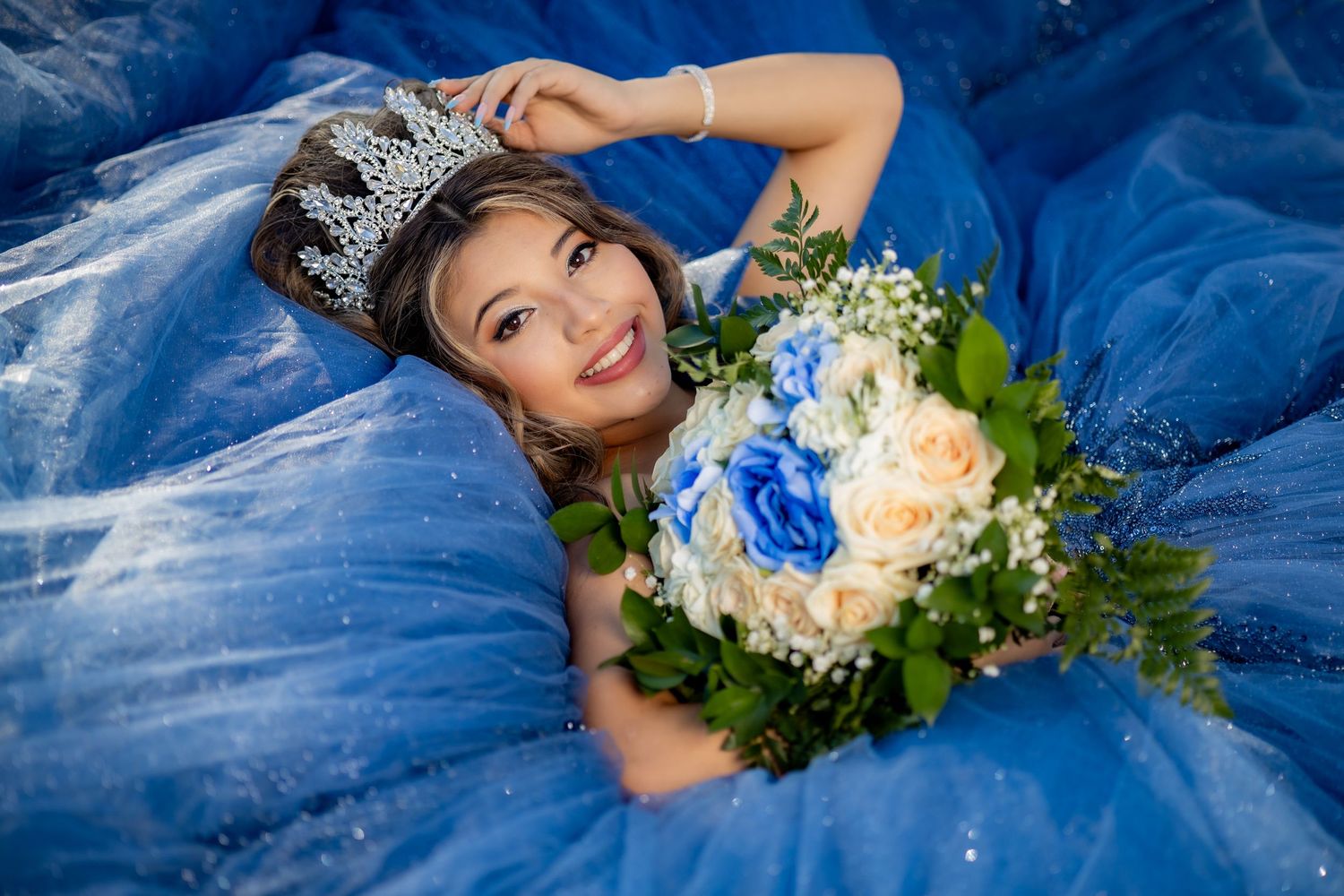 Smiling young woman in a sparkling blue quinceañera gown and silver tiara holds a bouquet of blue and peach roses with greenery.