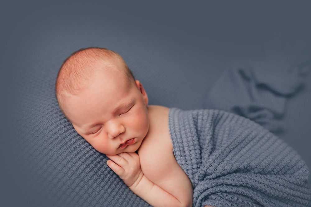 Professional newborn photography with baby wrapped in grey against dark blue background.