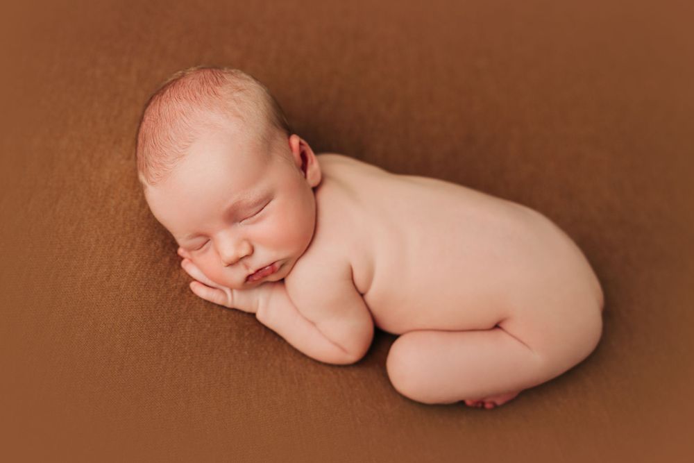 Artistic newborn photography with baby posed on warm brown backdrop.