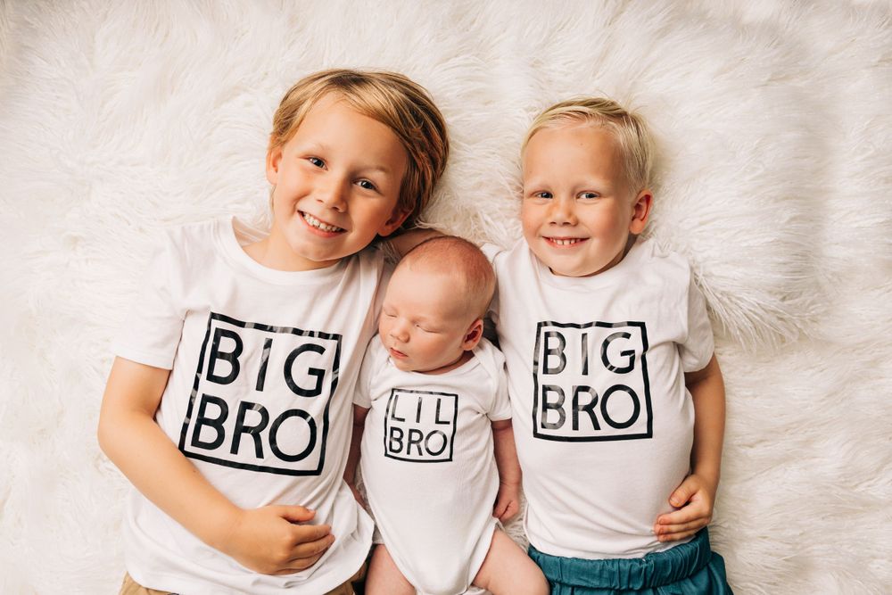 Three young children wearing matching 'BIG BRO' t-shirts pose together on a white fluffy rug.