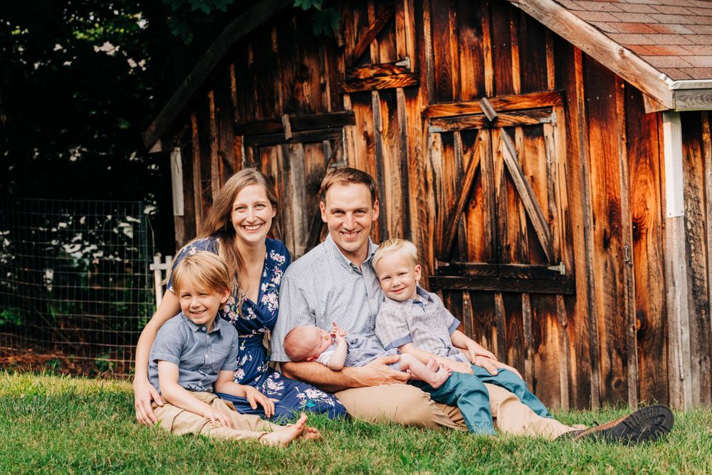 Family photo session outdoors against a rustic wooden barn backdrop in natural lighting.