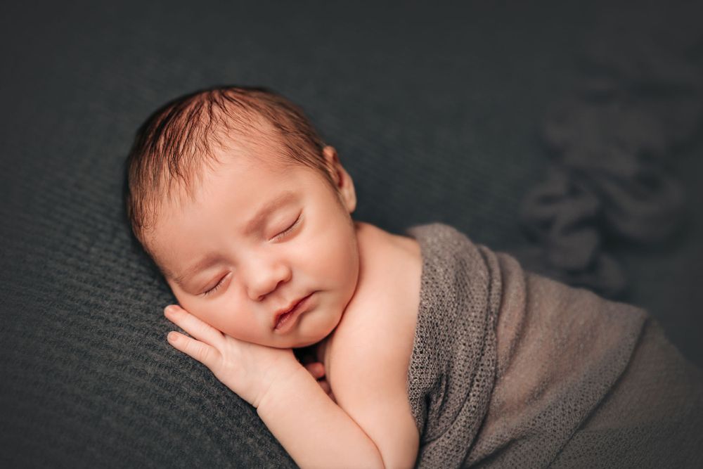 Sleeping newborn wrapped in gray swaddle against dark background in artistic portrait.