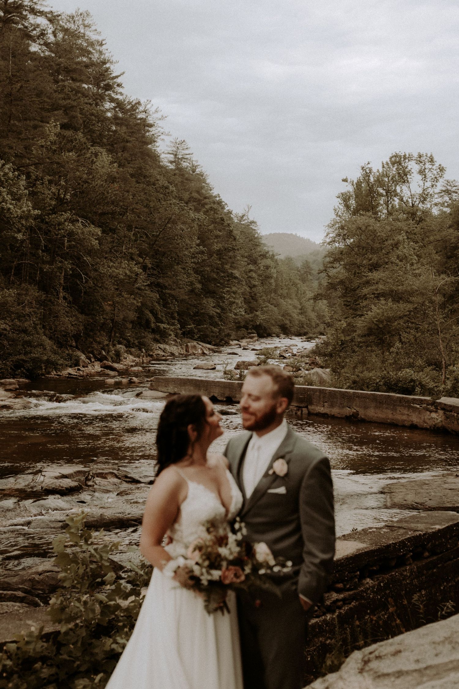 A romantic wedding portrait set against a flowing river and forest landscape.
