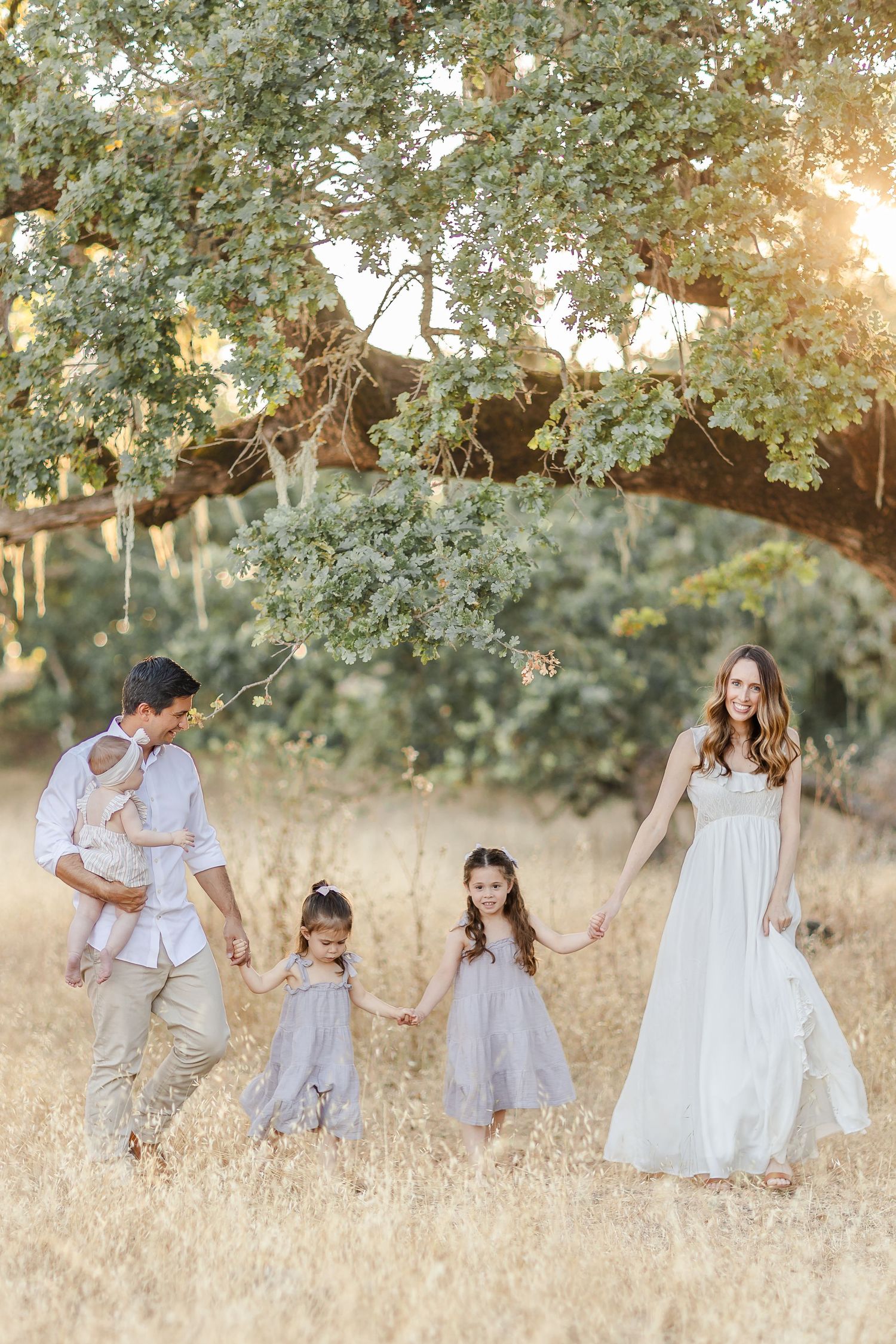 family of 2 parents and 3 girls in long dresses walking under a large tree at sunset and smiling. light and airy photographer Sonoma County.