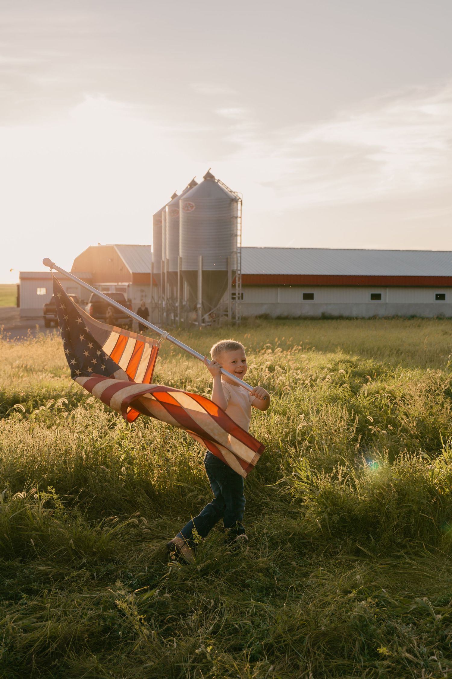 American flag waves in the golden sunlight on a farm with grain silos and barn buildings in the background.