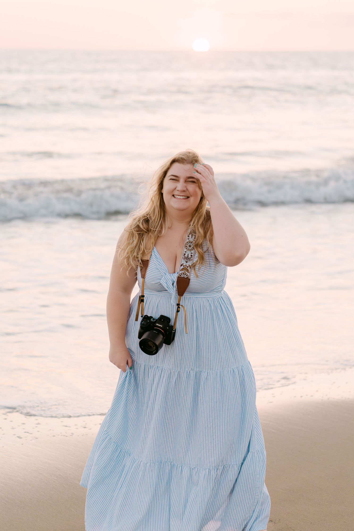 A person in a light blue tiered maxi dress poses on a sandy beach at sunset.