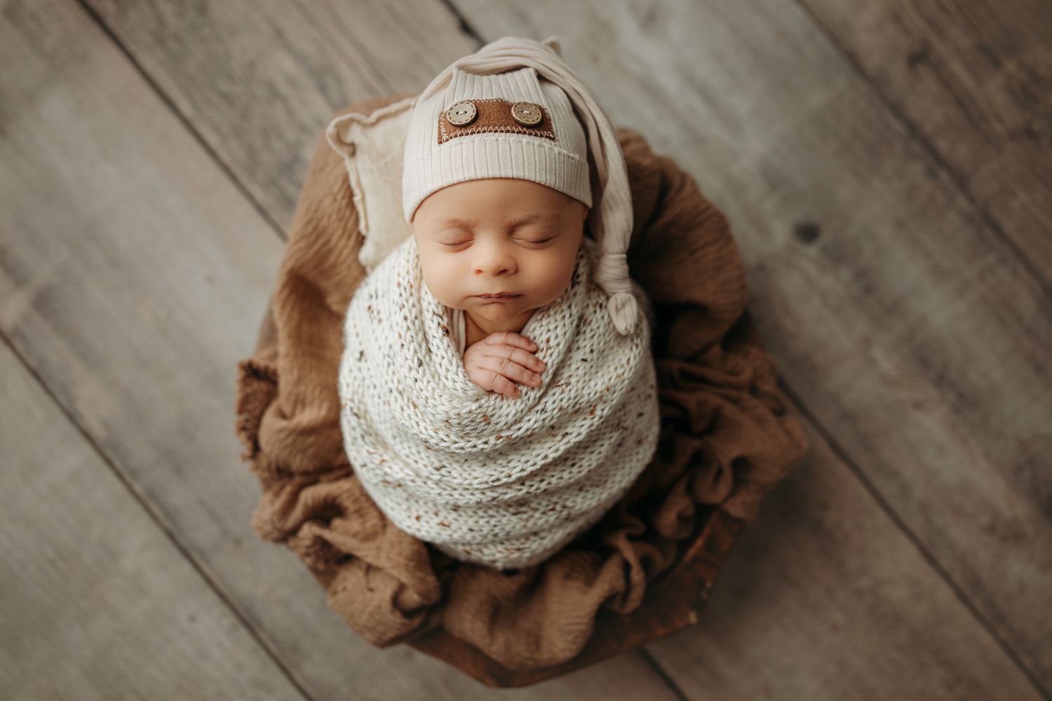 Sleeping newborn baby swaddled in cream knit blanket resting on brown fabric against wooden floor.