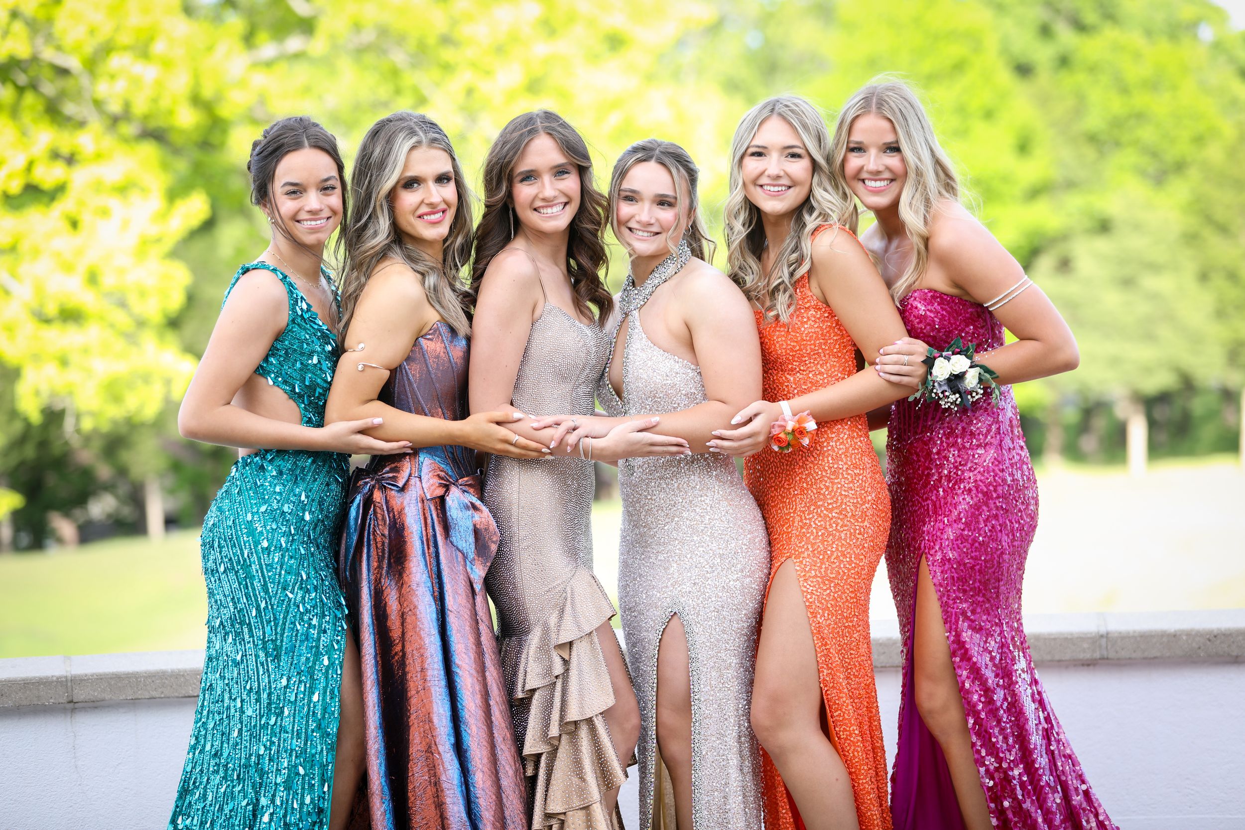 A group of girls in in colorful sequined evening gowns pose together outdoors in a sunny garden setting before heading to prom.