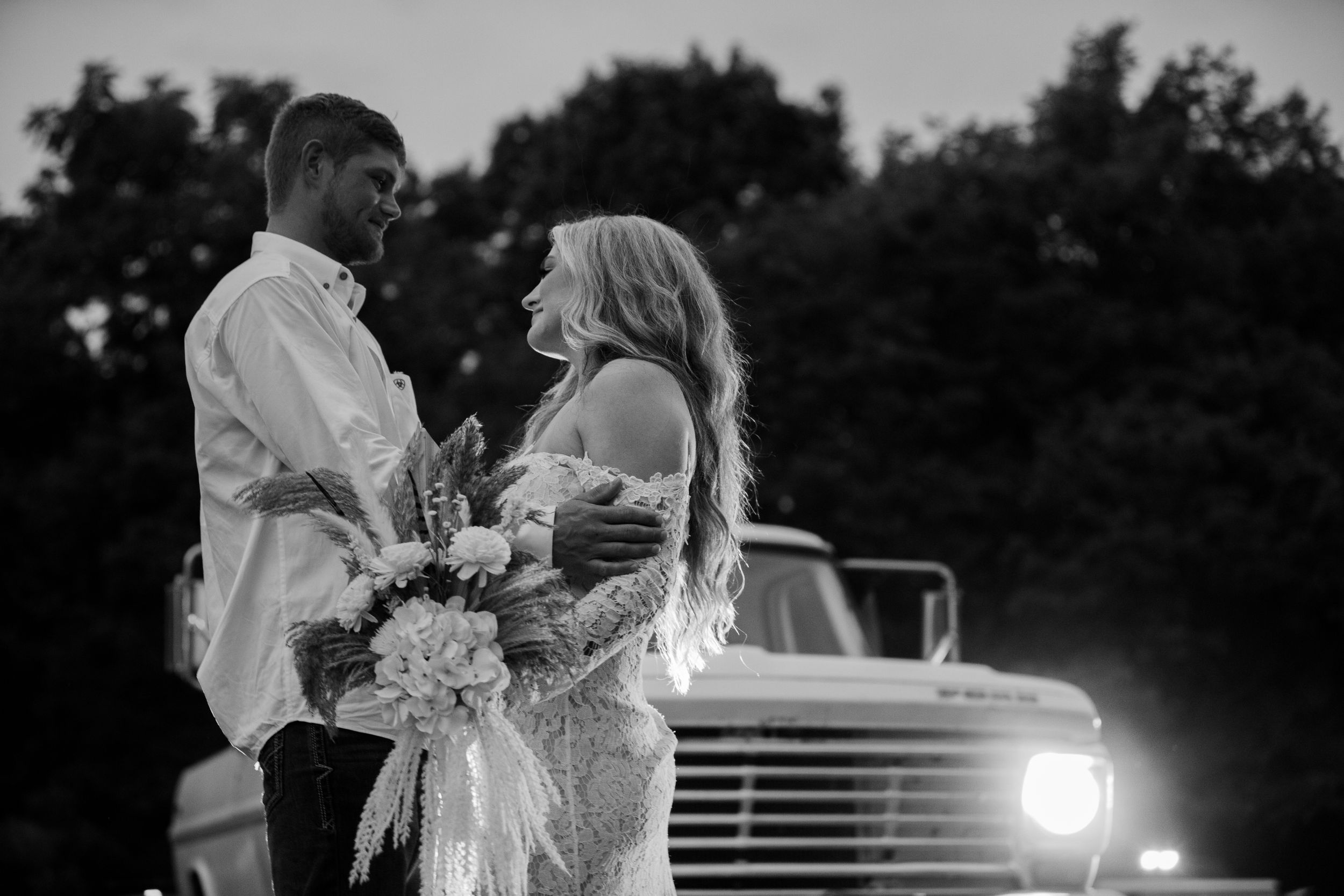 a bride and groom with a large bouquet in front of a vintage truck at dusk in black and white