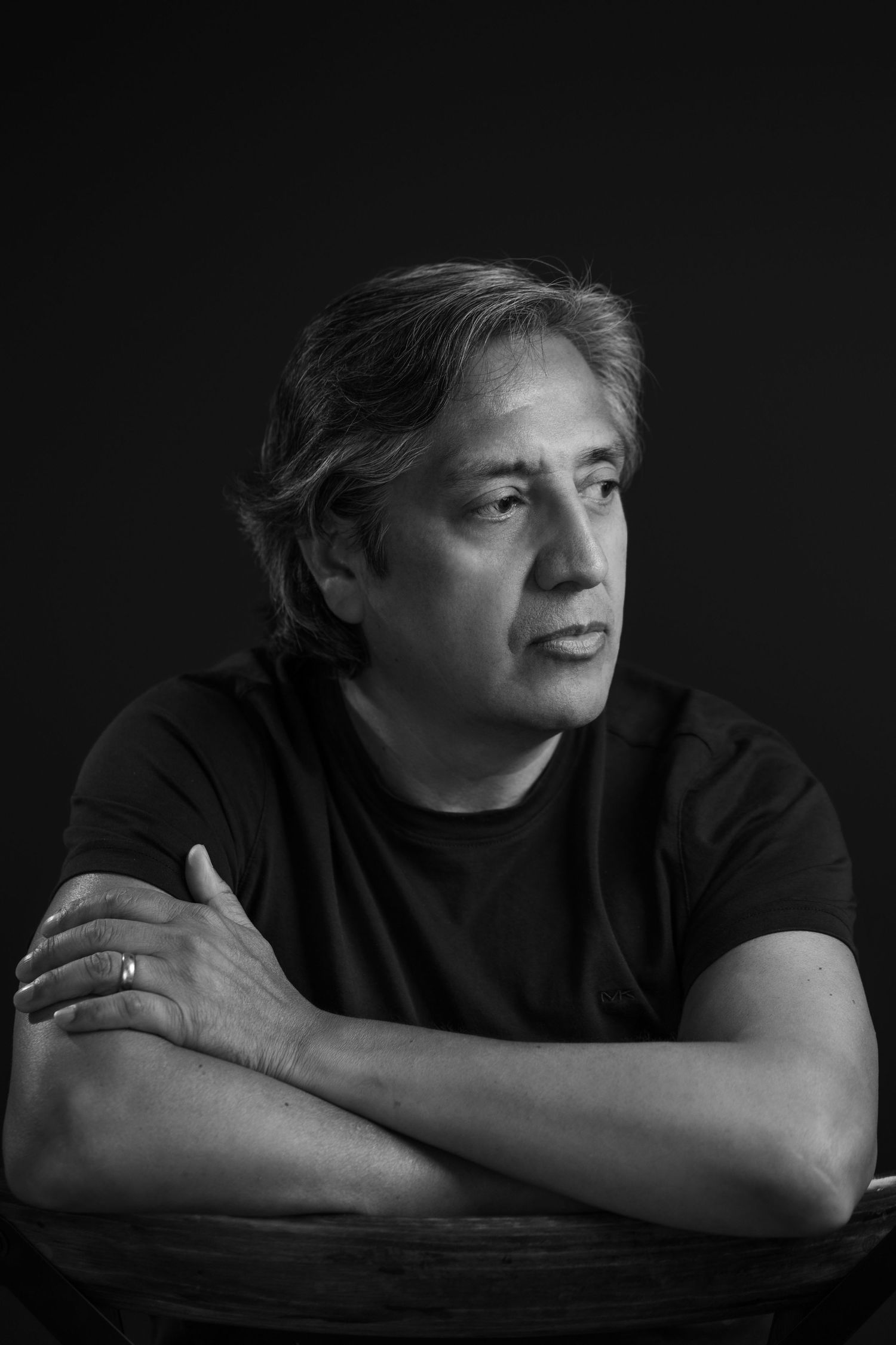 Black and white artistic portrait of a person in dark clothing against a black background with crossed arms and pensive expression.