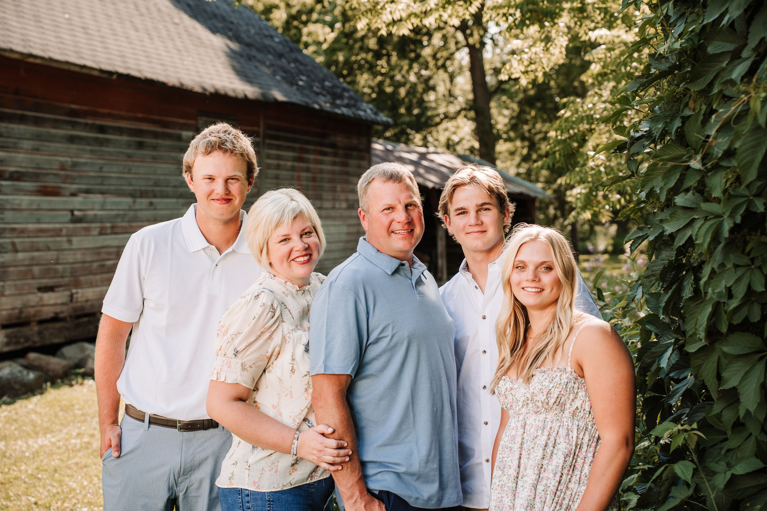 Happy family of five posing outdoors near a rustic wooden barn surrounded by lush green trees.