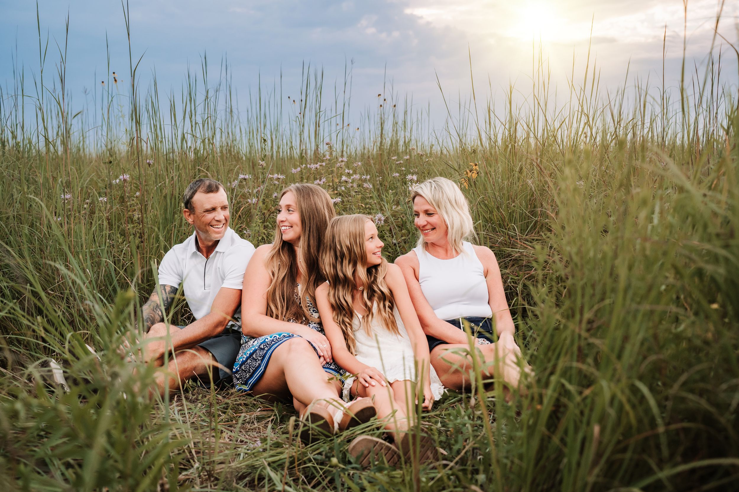 A family sits together amongst tall grass and sand dunes during sunset at the beach.