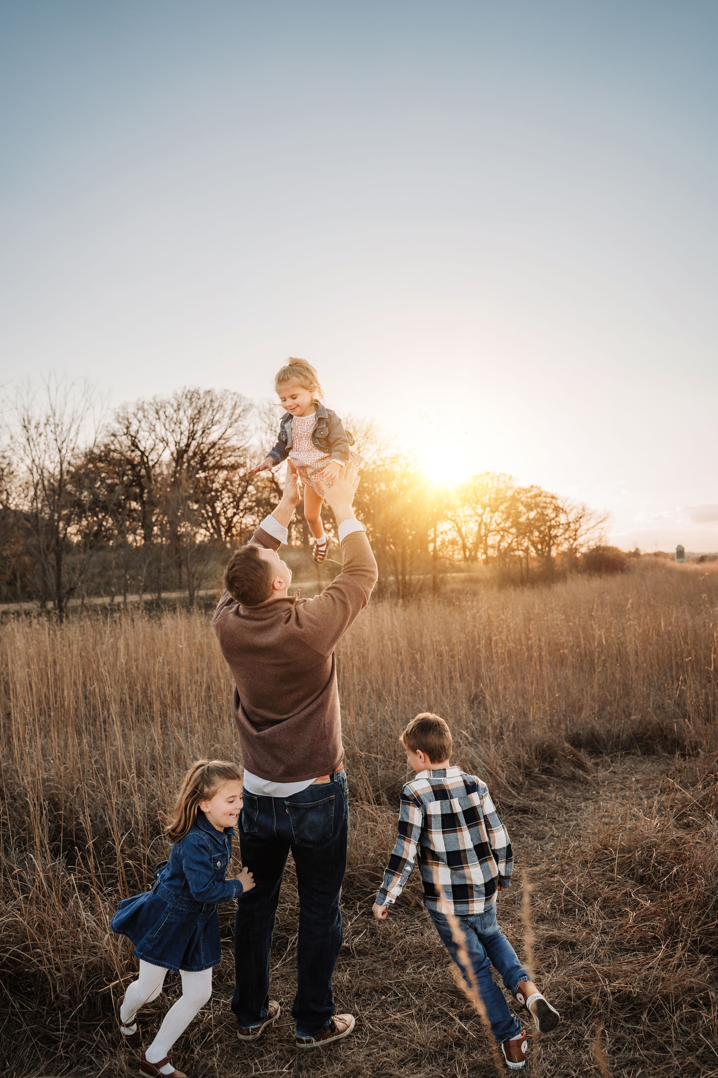 Family playing together in a golden field at sunset with tall grass and trees silhouetted in the background.