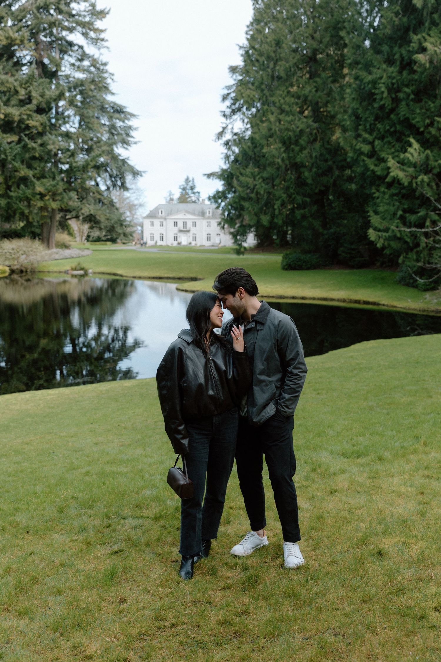 Couple in black jackets sharing a romantic moment on green lawn near a pond with a white estate in background.