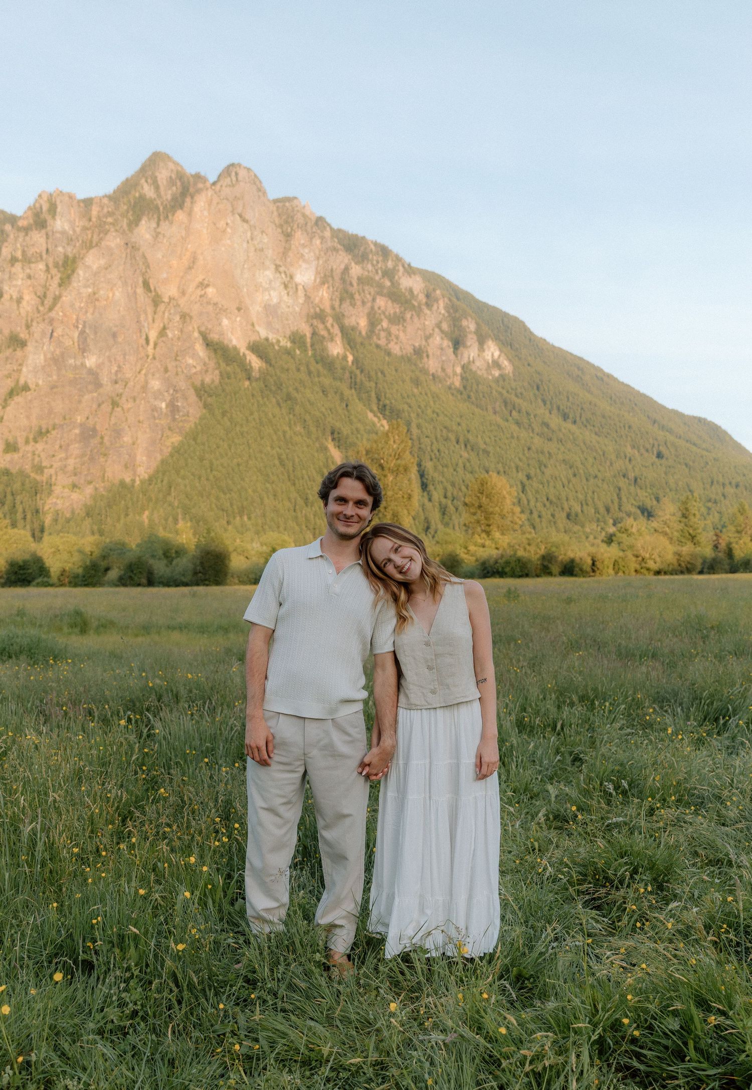 Couple standing in green meadow with dramatic mountain peaks in background during golden hour.