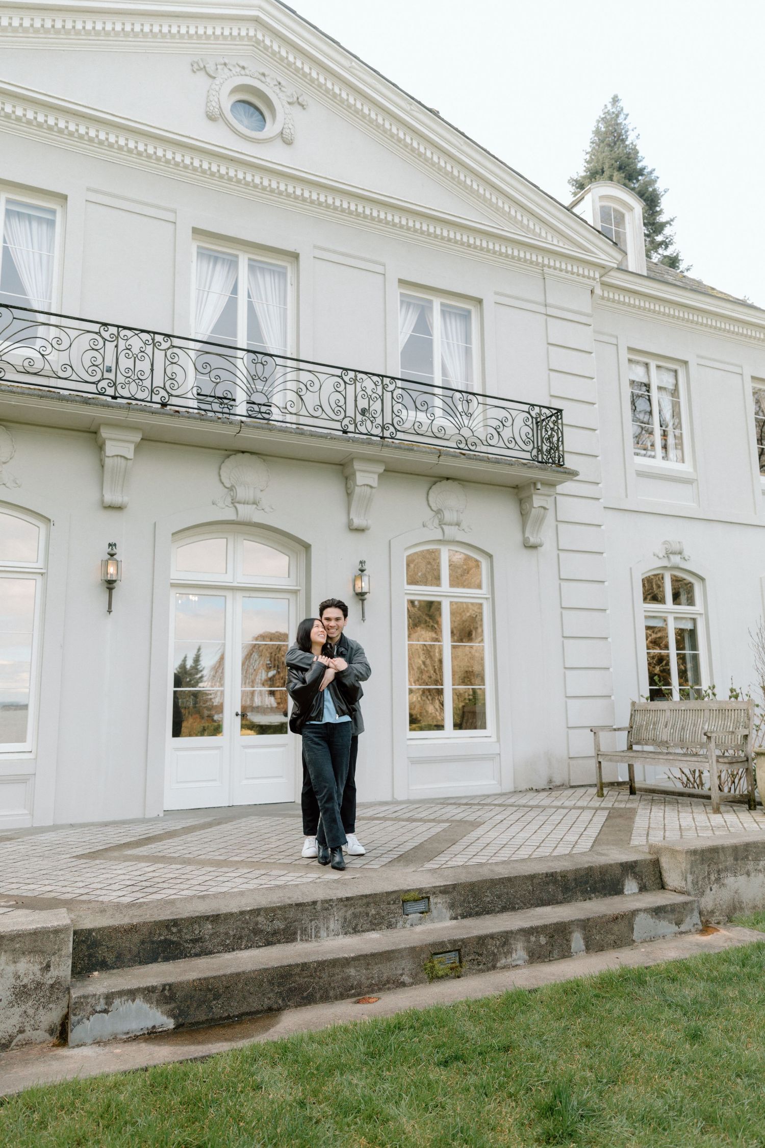 Couple embracing on steps of elegant white mansion with ornate balcony and French doors.