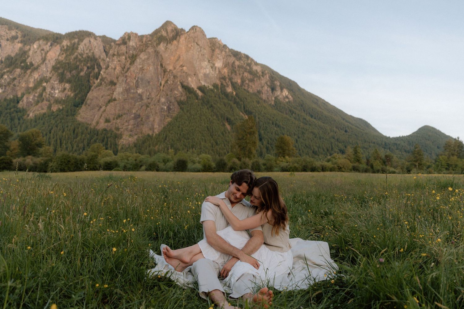 Couple sitting together in a green meadow with a dramatic mountain backdrop during golden hour.
