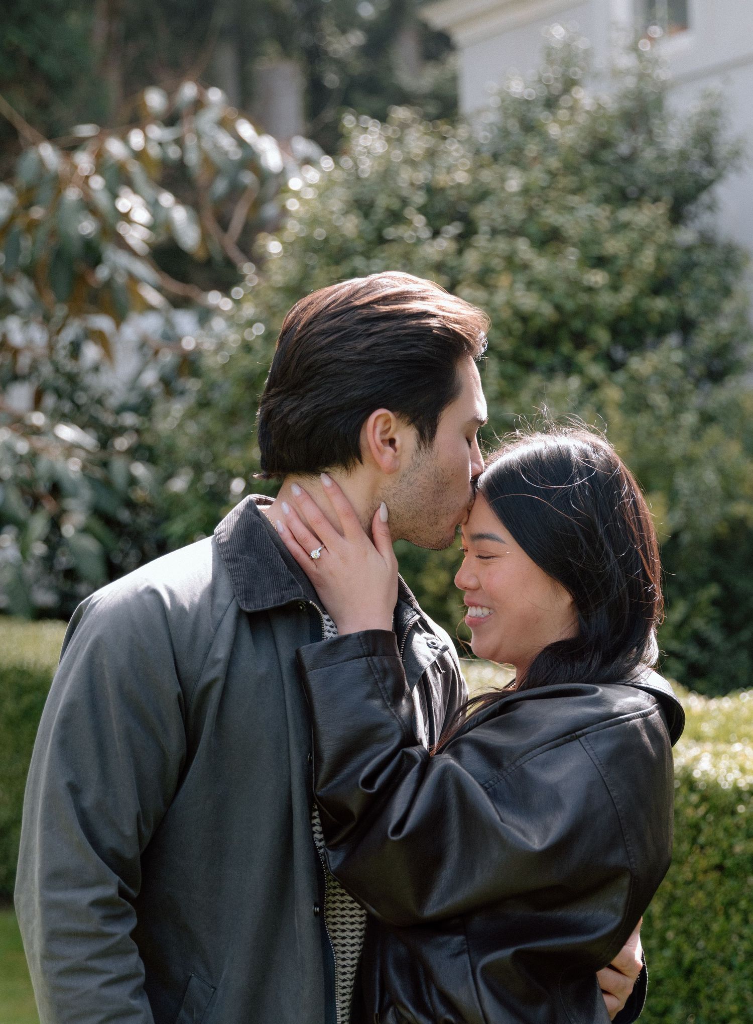 A couple shares a tender kiss outdoors, surrounded by lush greenery, both wearing leather jackets.