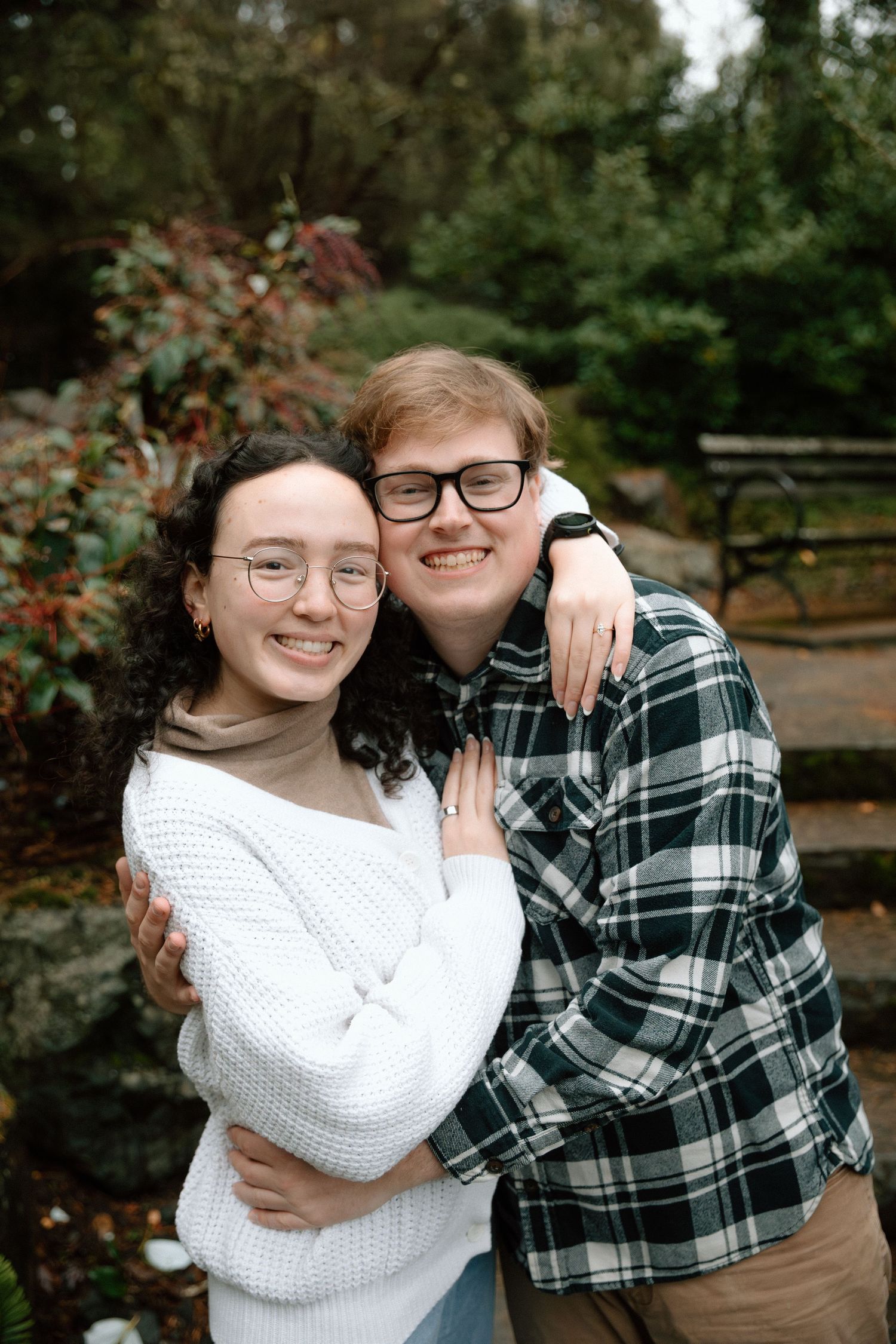 A couple wearing glasses embrace outdoors, with one in a white sweater and one in a plaid flannel shirt.