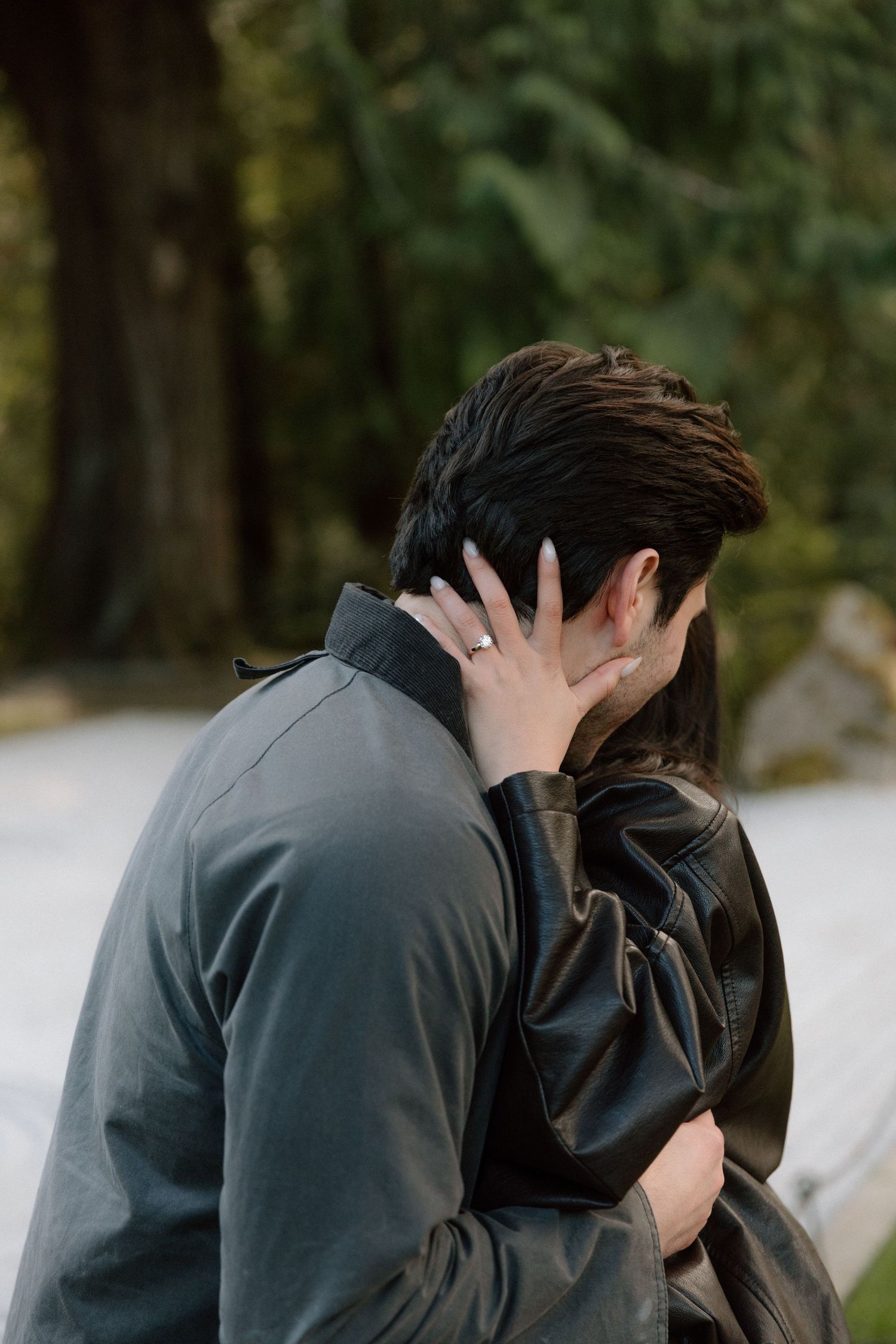 Couple embracing and kissing outdoors, woman wearing engagement ring, surrounded by lush green trees.