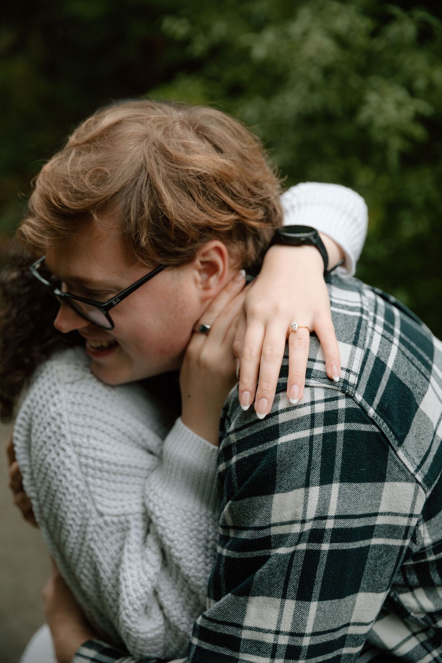 Couple embracing outdoors, woman wearing engagement ring hugs smiling man in plaid shirt with green trees behind.