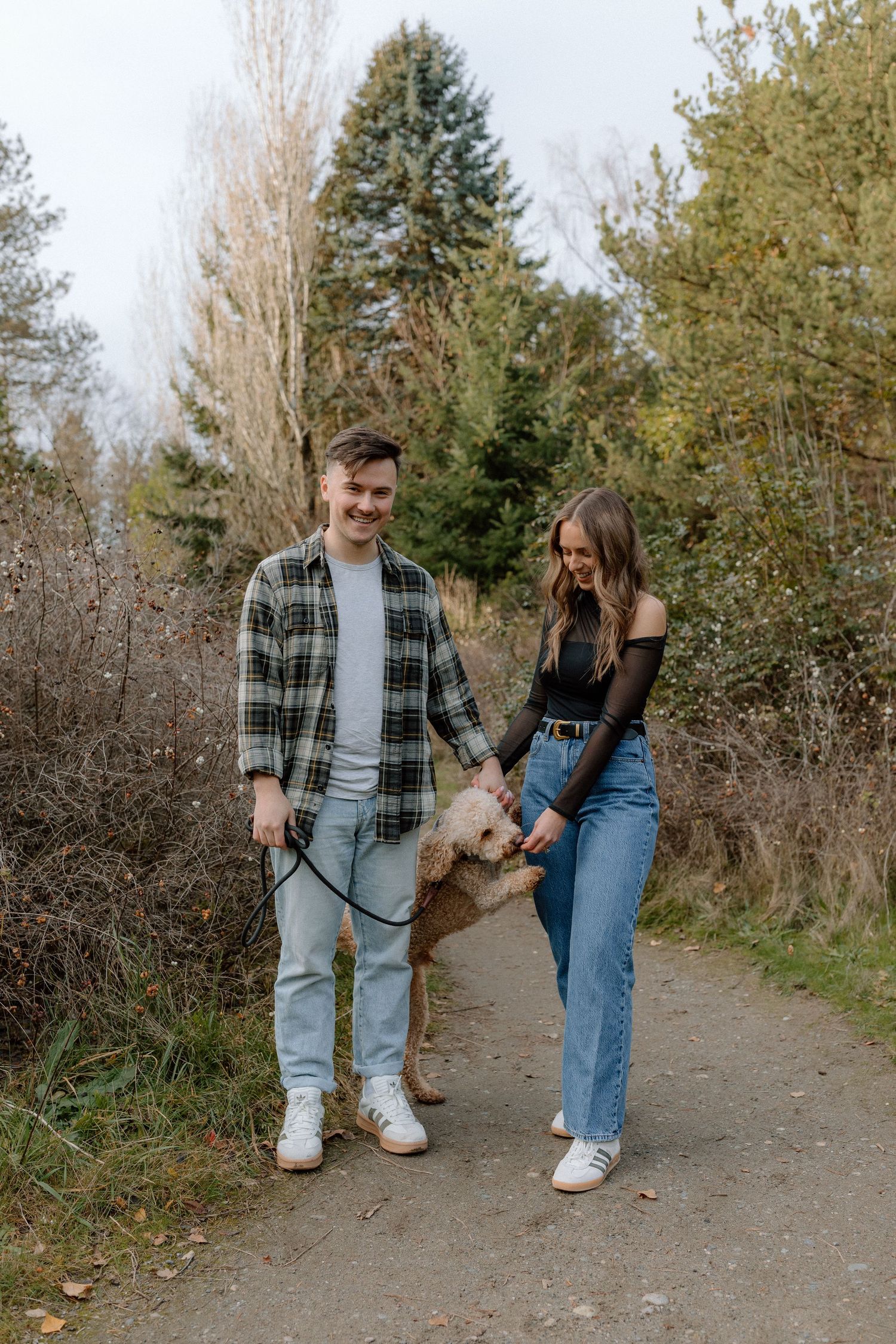 A smiling couple walks their golden doodle dog on a scenic nature path surrounded by lush green trees.