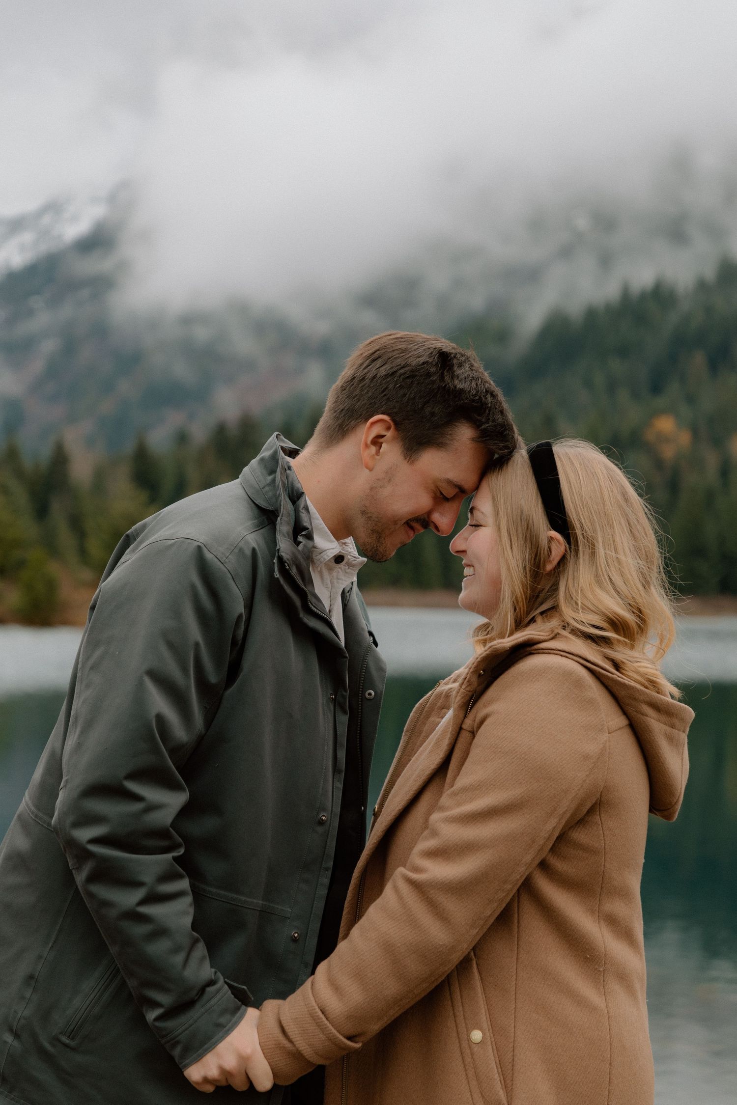 A couple in coats sharing an intimate moment in front of a misty mountain lake surrounded by evergreen forest.