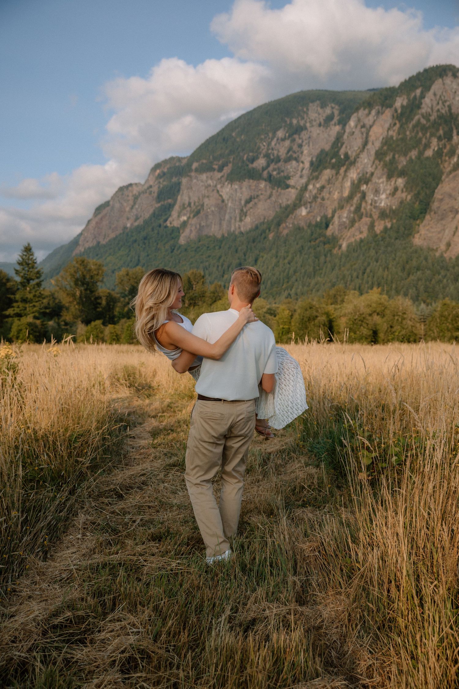 Couple embracing in golden field with mountain backdrop during sunset.