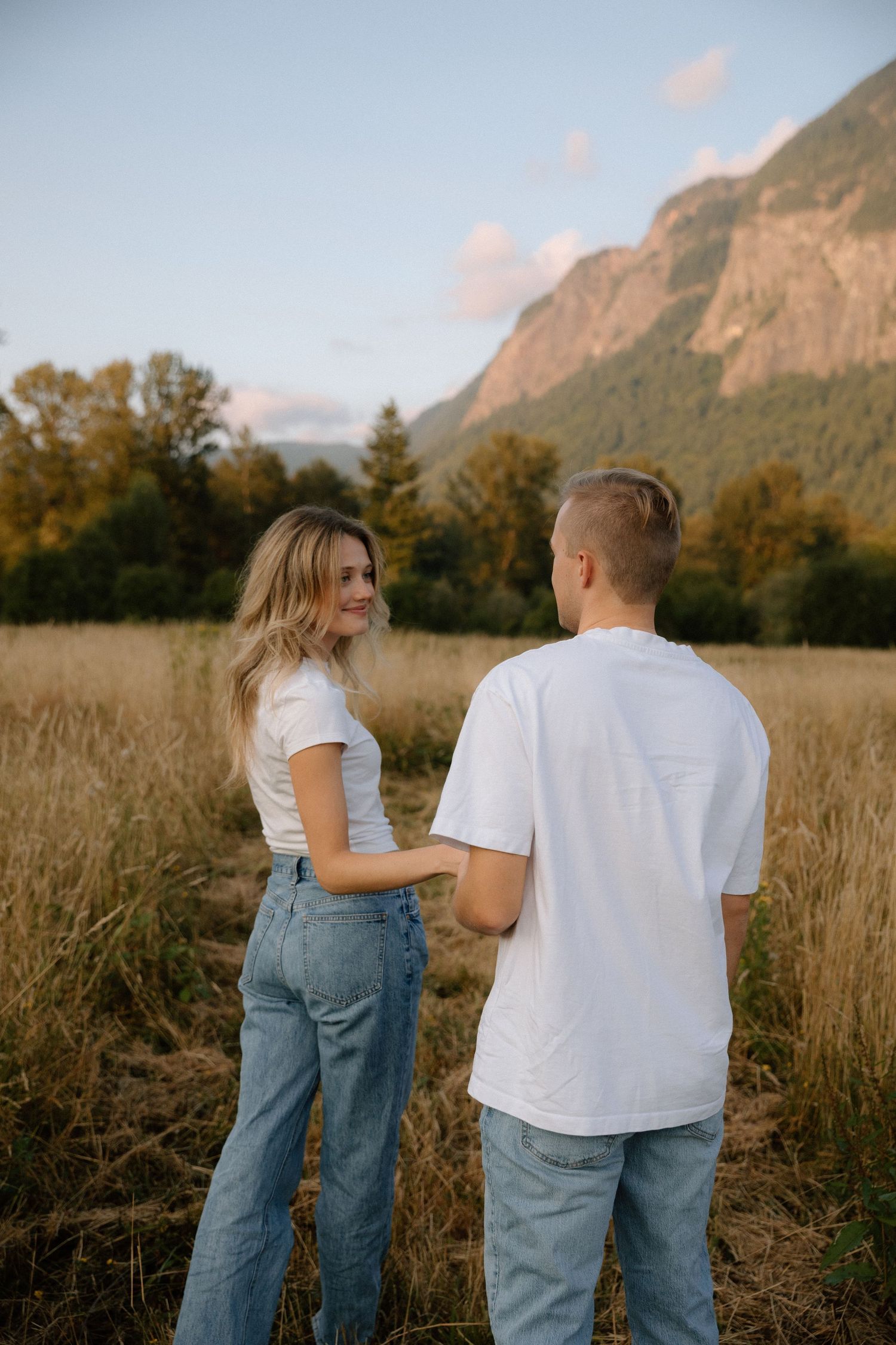 Couple holding hands in golden field with mountain backdrop at sunset, both wearing white tees and jeans.