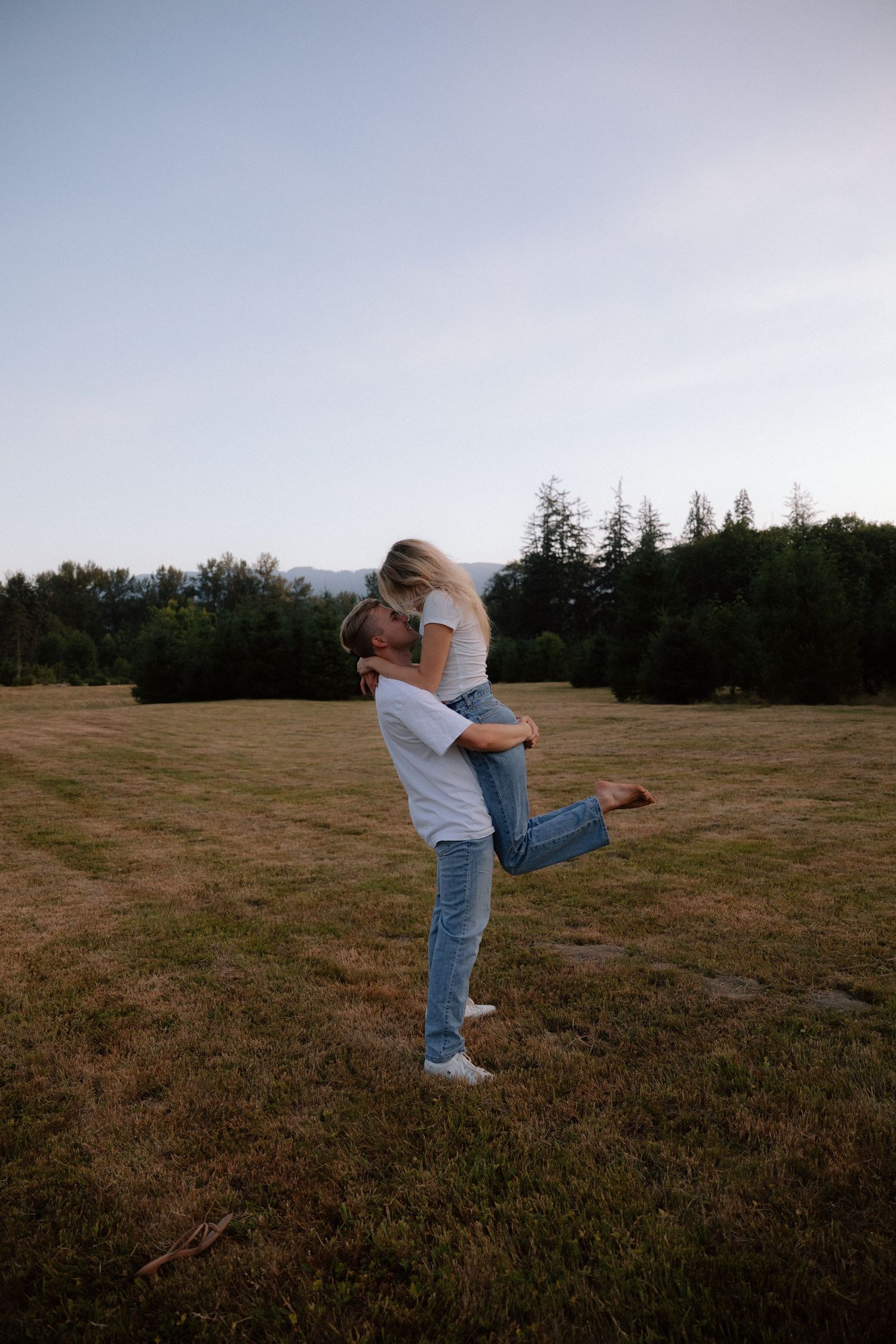 A couple embraces romantically in an open grassy field, with the man lifting the woman as they kiss at dusk.