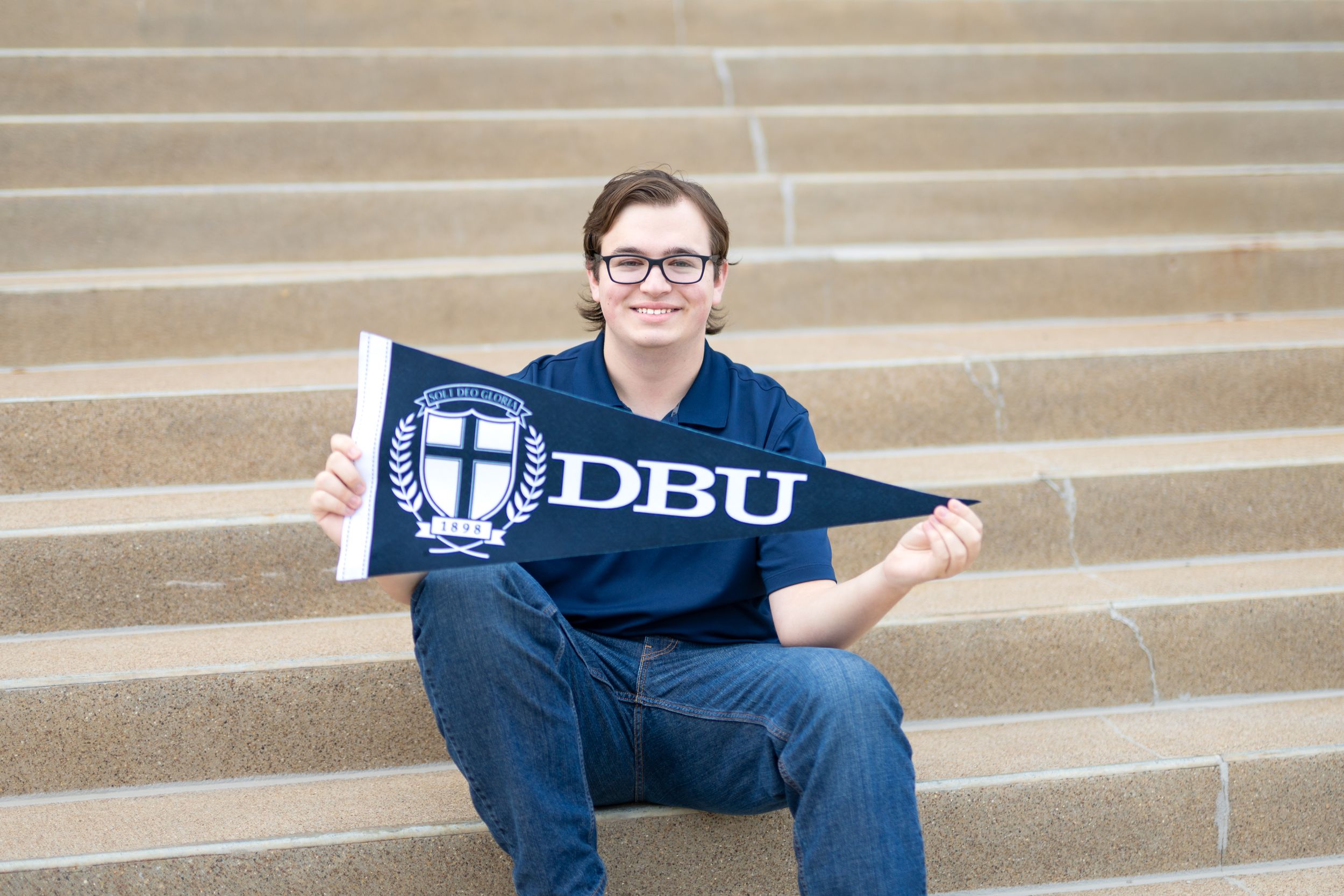 A student sits on concrete steps while holding a DBU university pennant.