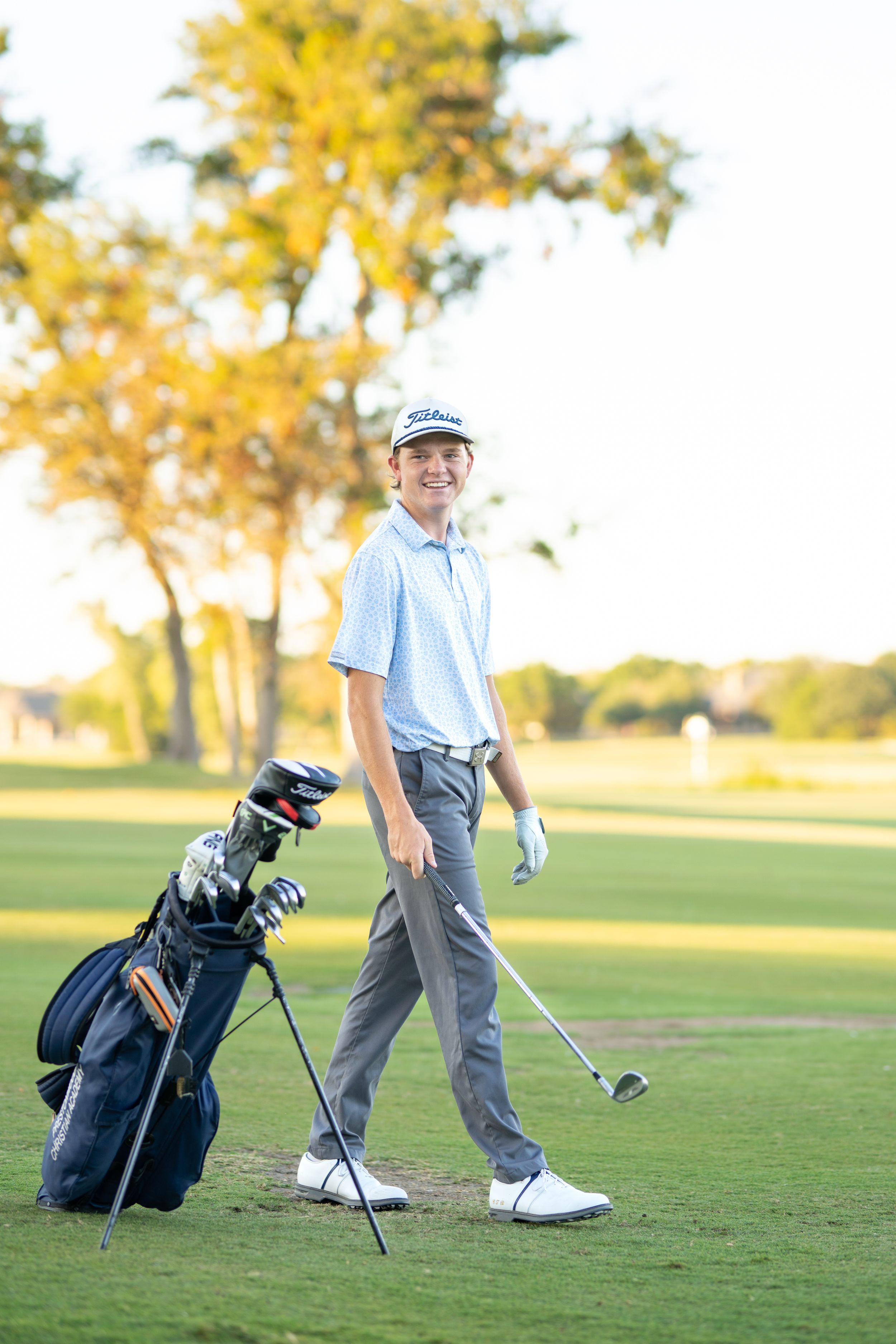 A golfer in blue polo and gray pants stands on a lush golf course with golf bag and clubs during autumn.