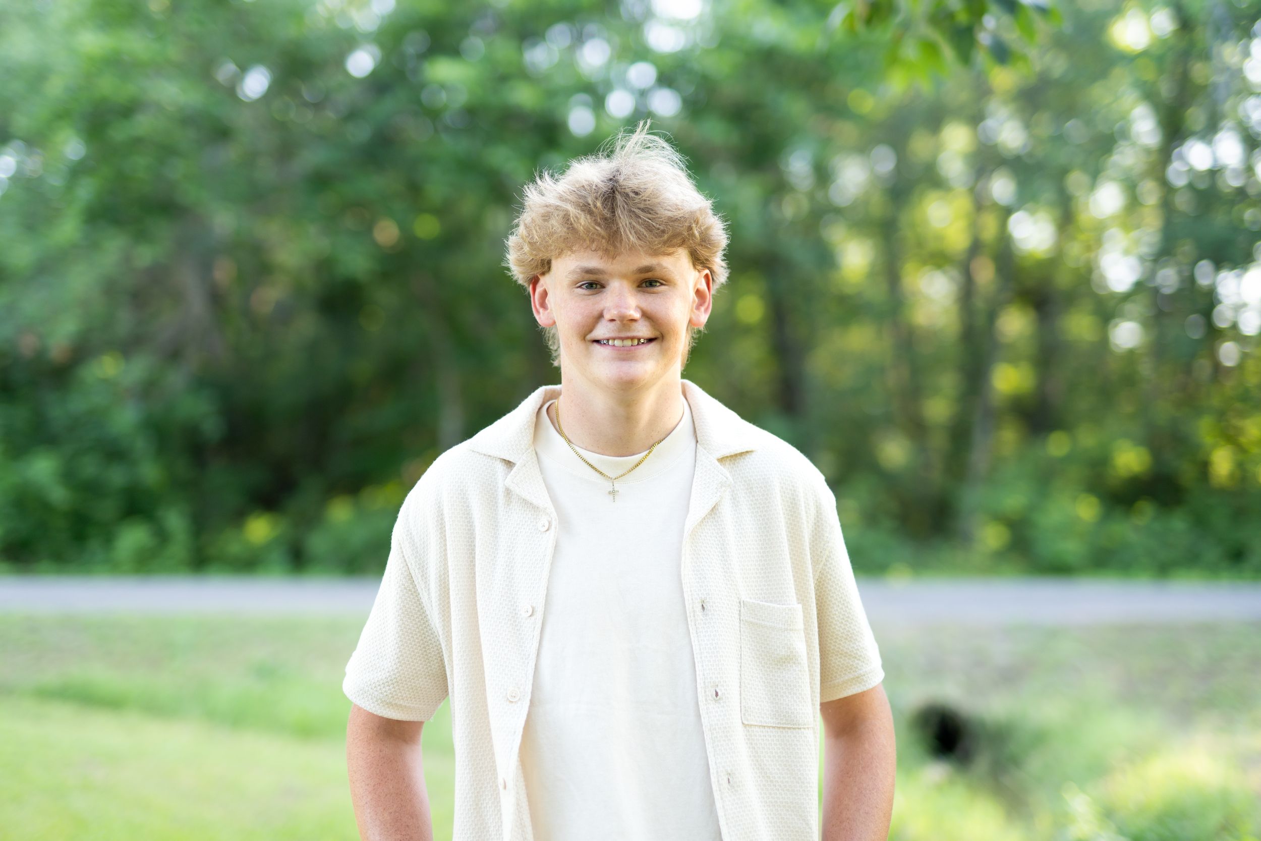 A casually dressed person stands outdoors on a sunny day with green foliage in the background.