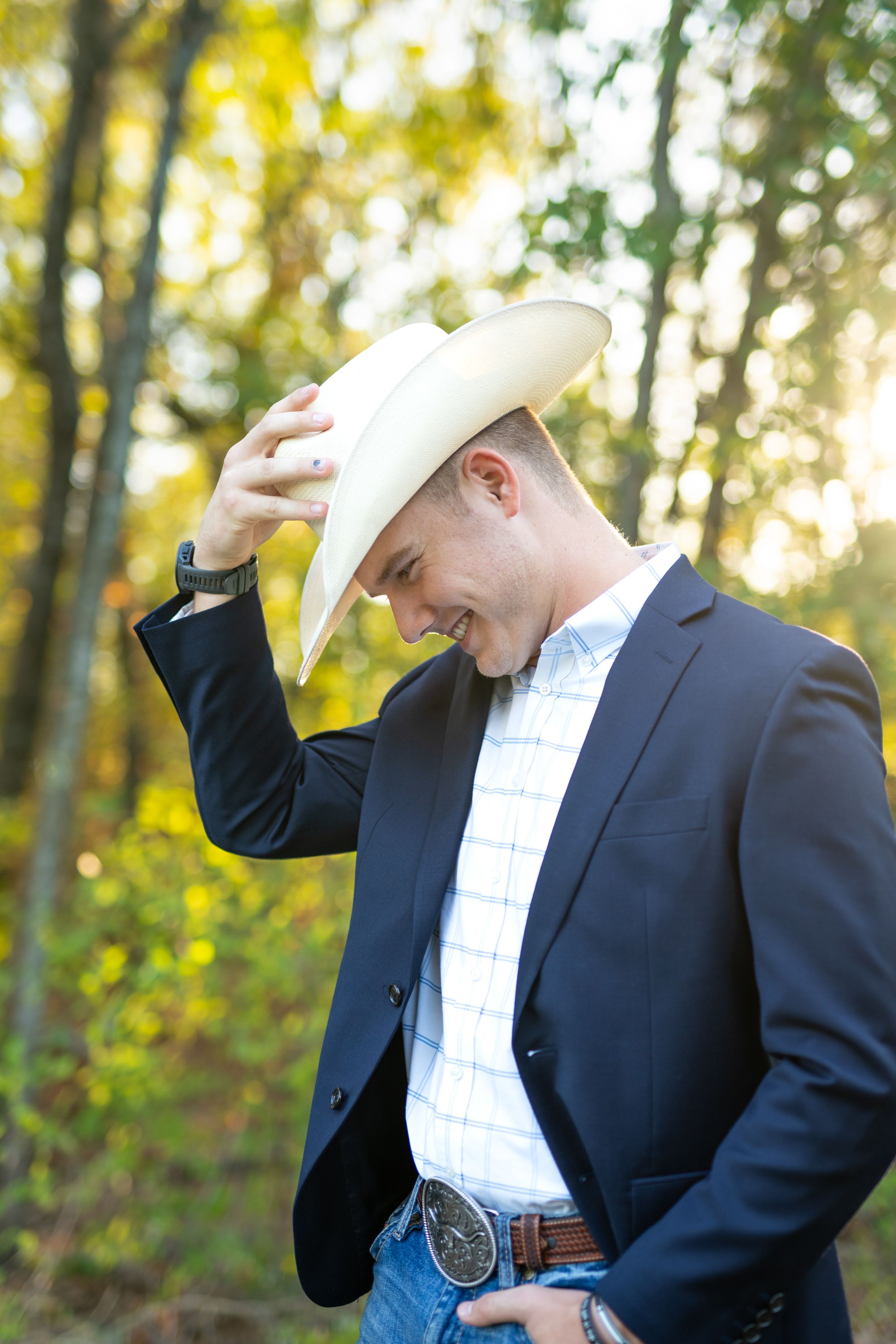 Man in a navy suit jacket and cowboy hat looks down while adjusting his hat against a blurred autumn forest background.