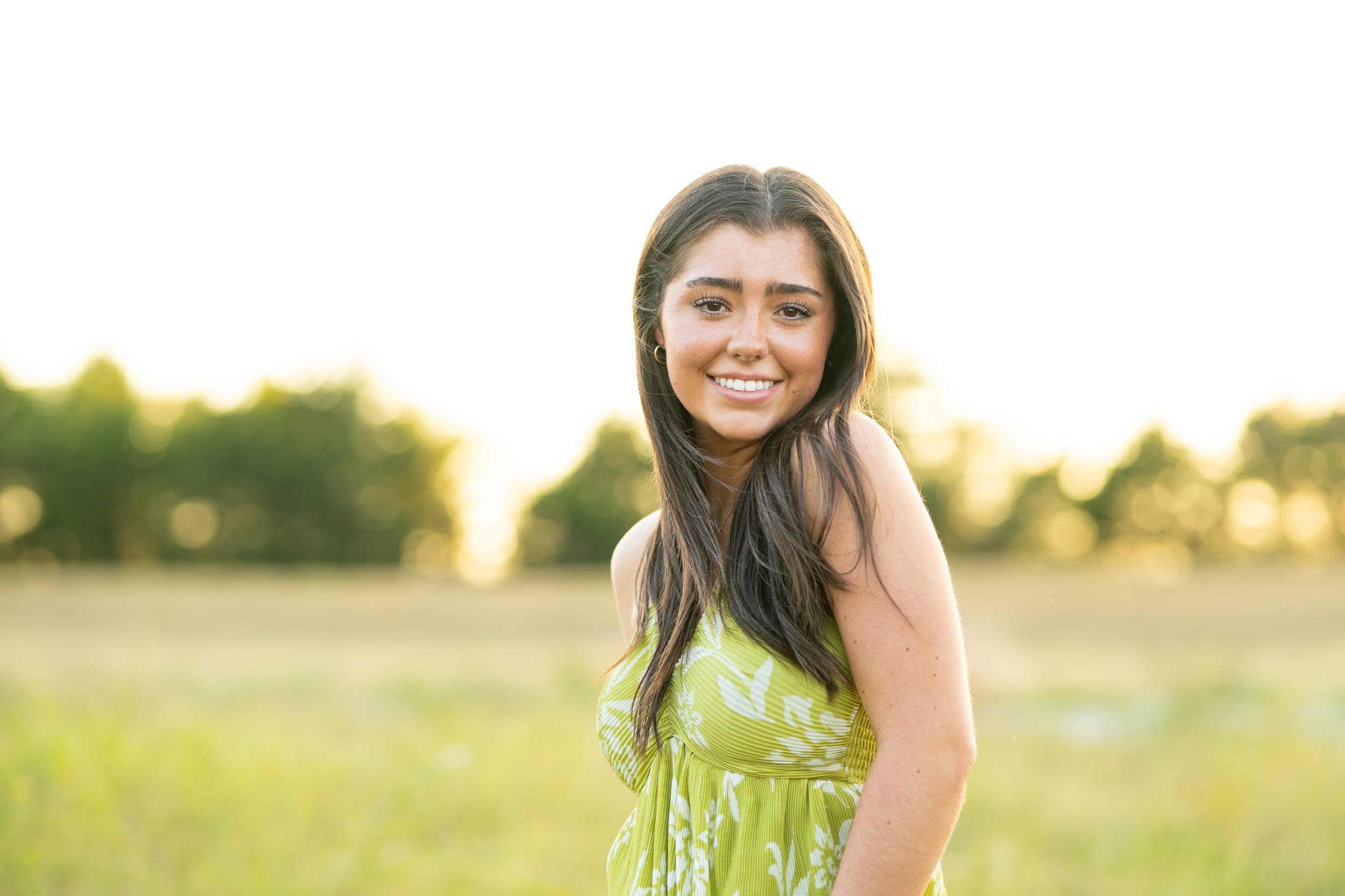 A smiling person in a green sundress poses in a sunlit meadow during golden hour.