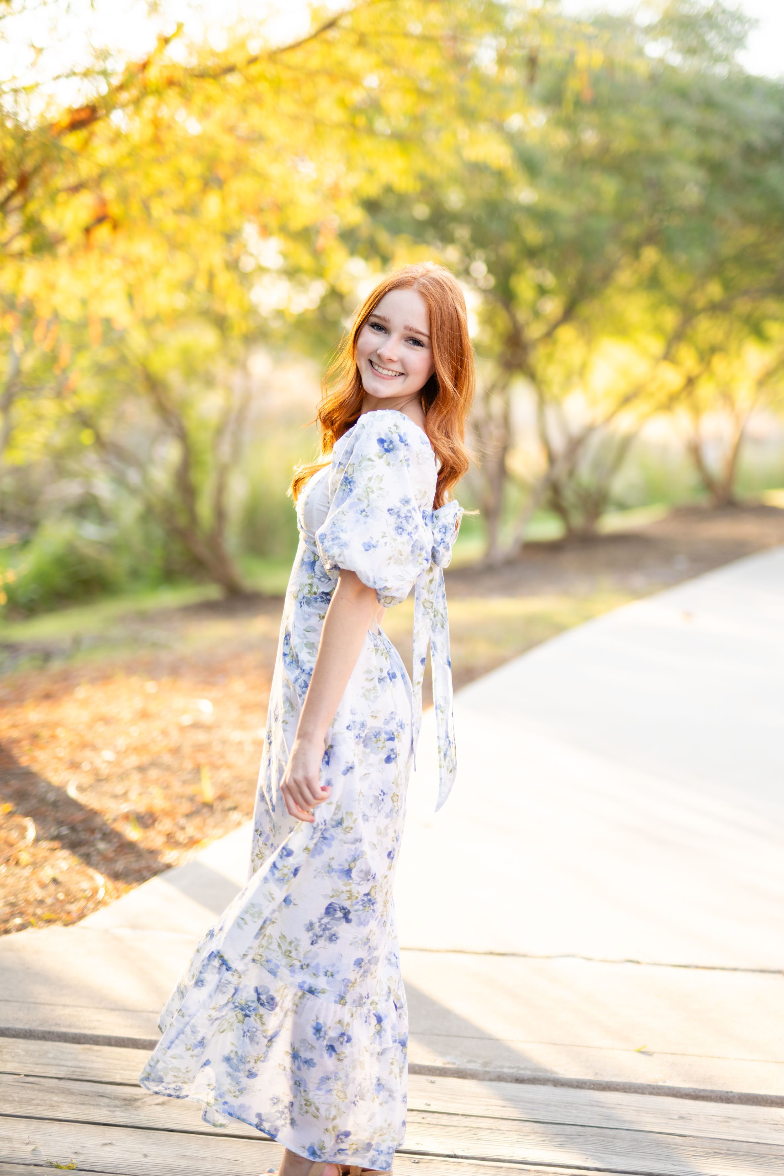 Someone in a flowing blue and white floral maxi dress poses on a pathway with golden autumn foliage in the background.