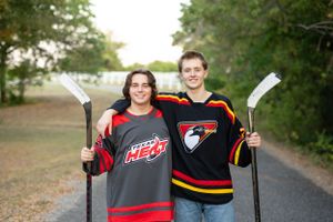 Two people in hockey jerseys stand on a path holding hockey sticks while smiling at the camera.