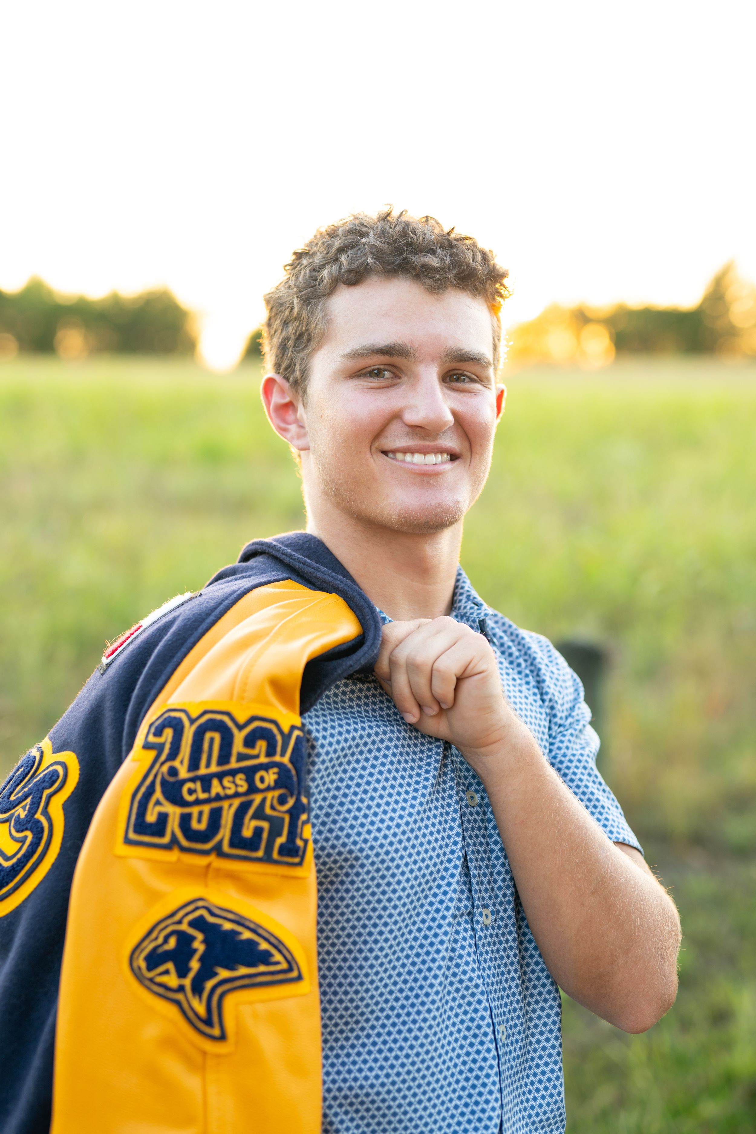 Student in a yellow and blue class of 2022 varsity letterman jacket smiles while standing outdoors at sunset.