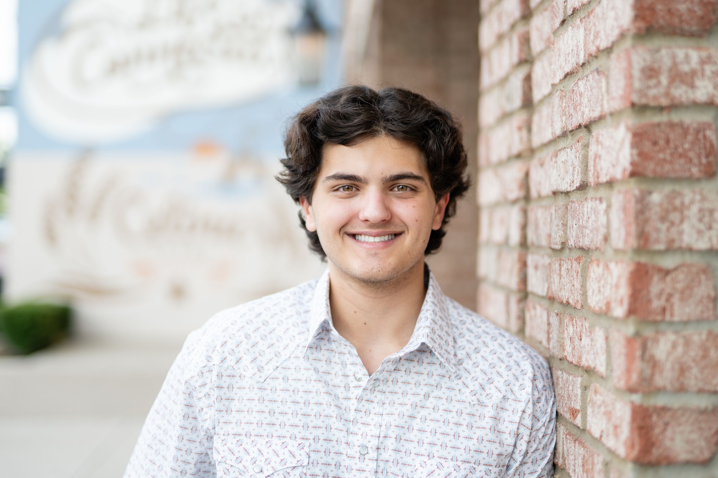 Professional headshot portrait of someone smiling in front of a brick wall wearing a patterned button down shirt.