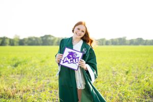 Graduate in green cap and gown stands in sunny field holding Eagle university sign and smiling.