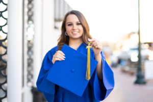 Graduate in bright blue cap and gown holds gold tassel while posing outdoors during graduation photoshoot.