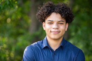 A smiling person with curly hair wearing a blue polka dot button-up shirt poses outdoors against a blurred green background.