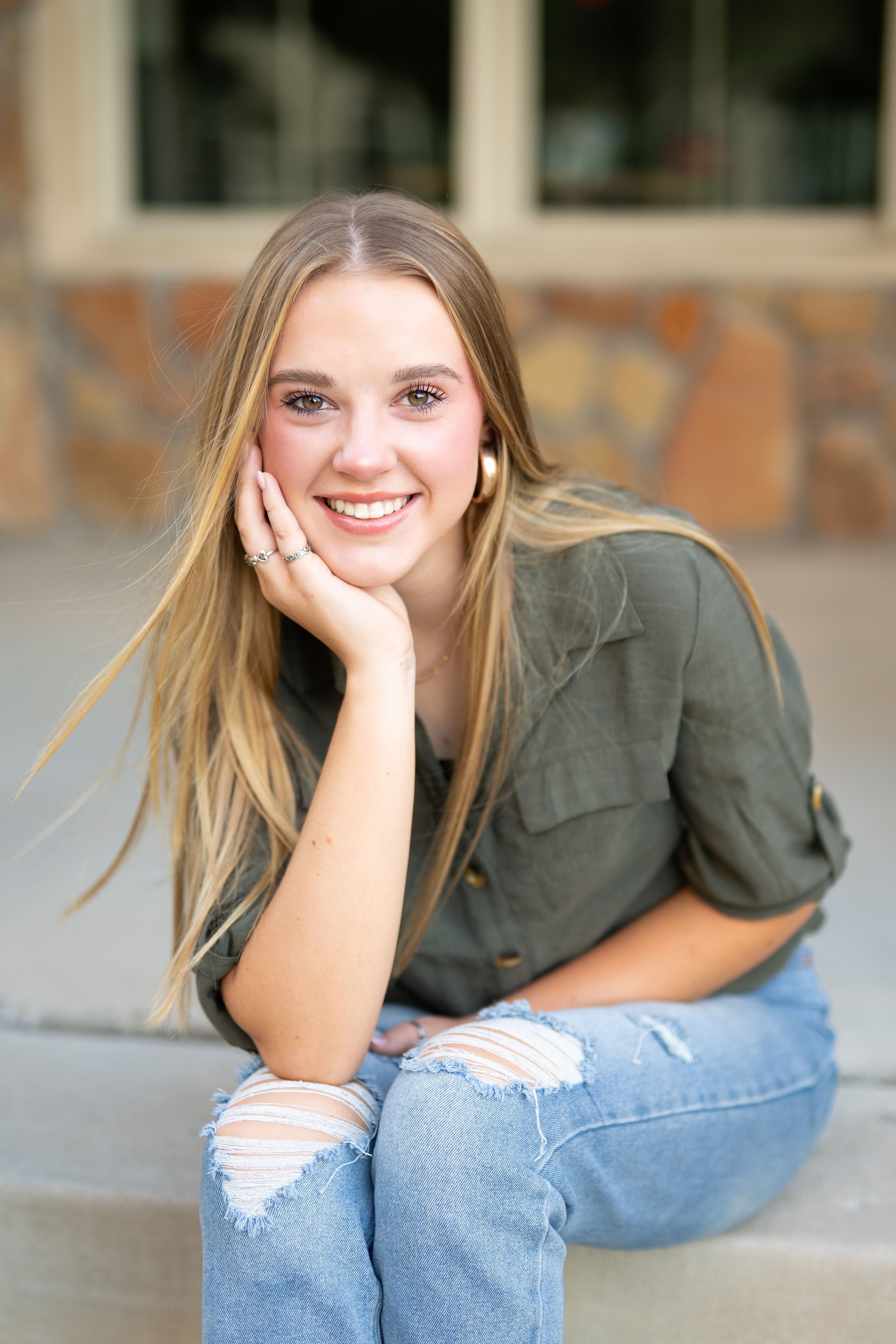 Young woman in olive button-down and ripped jeans sits casually on porch with hand resting on chin and bright smile.