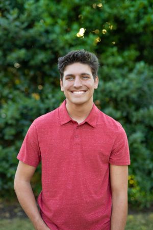 Someone wearing a red polo shirt smiles while standing outdoors with greenery in the background.