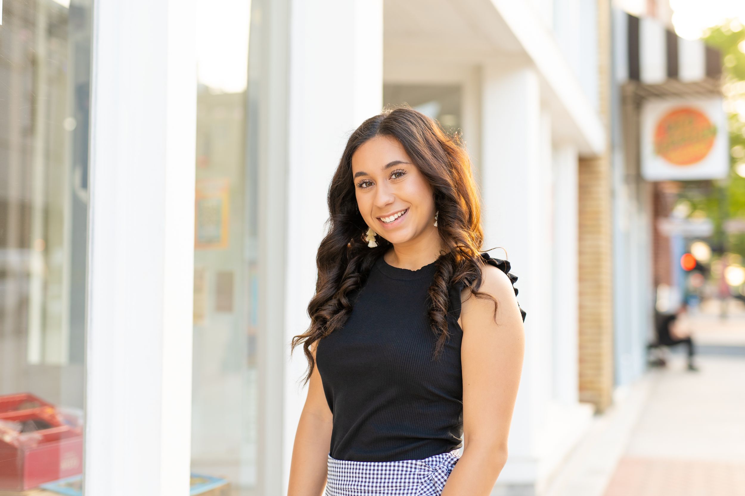 A person wearing a black sleeveless top and gingham pants stands smiling on an urban sidewalk.