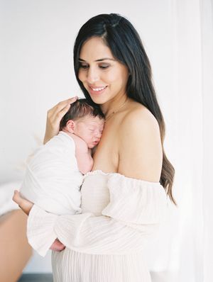 A tender moment captured in soft light as a mother in white holds her sleeping newborn baby against her chest.